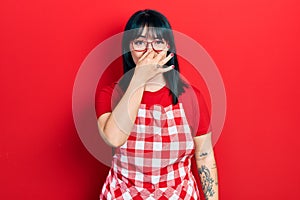 Young hispanic woman wearing cook apron and glasses smelling something stinky and disgusting, intolerable smell, holding breath