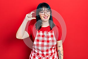 Young hispanic woman wearing cook apron and glasses smiling and confident gesturing with hand doing small size sign with fingers