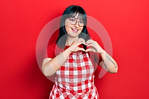 Young hispanic woman wearing cook apron and glasses smiling in love doing heart symbol shape with hands