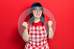 Young hispanic woman wearing cook apron and glasses very happy and excited doing winner gesture with arms raised, smiling and