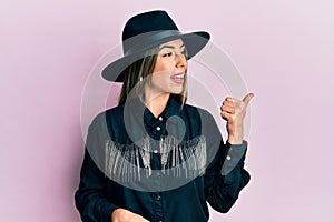 Young hispanic woman wearing cowgirl style smiling with happy face looking and pointing to the side with thumb up