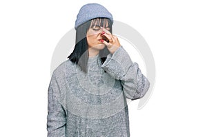 Young hispanic woman wearing cute wool cap smelling something stinky and disgusting, intolerable smell, holding breath with