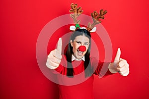 Young hispanic woman wearing deer christmas hat and red nose approving doing positive gesture with hand, thumbs up smiling and