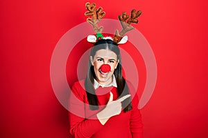 Young hispanic woman wearing deer christmas hat and red nose cheerful with a smile on face pointing with hand and finger up to the