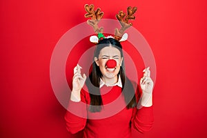 Young hispanic woman wearing deer christmas hat and red nose gesturing finger crossed smiling with hope and eyes closed