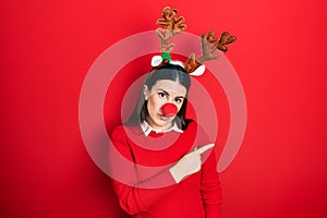 Young hispanic woman wearing deer christmas hat and red nose pointing with hand finger to the side showing advertisement, serious