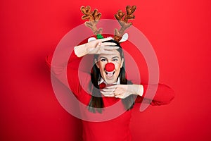 Young hispanic woman wearing deer christmas hat and red nose smiling cheerful playing peek a boo with hands showing face