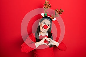 Young hispanic woman wearing deer christmas hat and red nose smiling in love doing heart symbol shape with hands