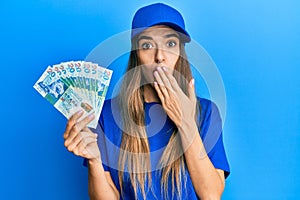 Young hispanic woman wearing delivery uniform and cap holding 20 hong kong dollars covering mouth with hand, shocked and afraid