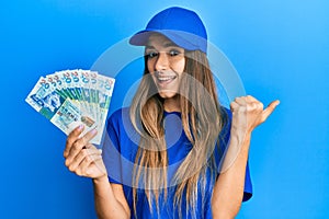 Young hispanic woman wearing delivery uniform and cap holding 20 hong kong dollars pointing thumb up to the side smiling happy