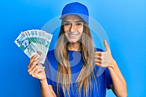 Young hispanic woman wearing delivery uniform and cap holding 20 hong kong dollars smiling happy and positive, thumb up doing