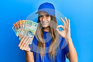 Young hispanic woman wearing delivery uniform and cap holding australian dollars doing ok sign with fingers, smiling friendly