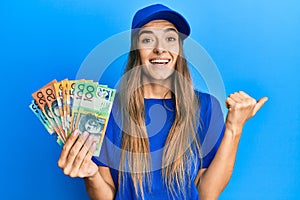 Young hispanic woman wearing delivery uniform and cap holding australian dollars pointing thumb up to the side smiling happy with