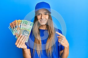 Young hispanic woman wearing delivery uniform and cap holding australian dollars smiling happy pointing with hand and finger