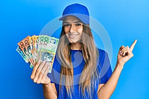 Young hispanic woman wearing delivery uniform and cap holding australian dollars smiling happy pointing with hand and finger to