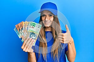 Young hispanic woman wearing delivery uniform and cap holding australian dollars smiling happy and positive, thumb up doing