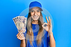 Young hispanic woman wearing delivery uniform and cap holding japanese yuan doing ok sign with fingers, smiling friendly gesturing