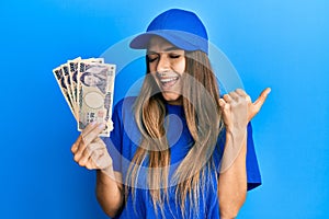 Young hispanic woman wearing delivery uniform and cap holding japanese yuan pointing thumb up to the side smiling happy with open