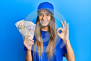 Young hispanic woman wearing delivery uniform and cap holding singapore dollars doing ok sign with fingers, smiling friendly