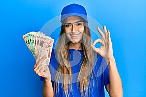 Young hispanic woman wearing delivery uniform and cap holding south korean won doing ok sign with fingers, smiling friendly