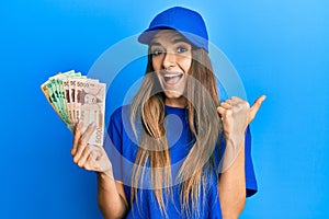 Young hispanic woman wearing delivery uniform and cap holding south korean won pointing thumb up to the side smiling happy with