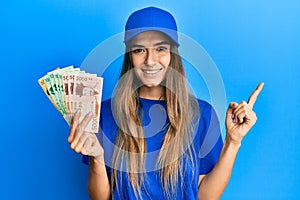 Young hispanic woman wearing delivery uniform and cap holding south korean won smiling happy pointing with hand and finger to the