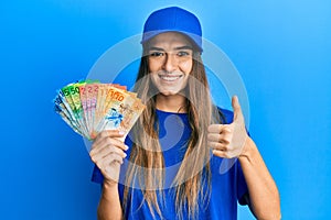 Young hispanic woman wearing delivery uniform and cap holding swiss franc smiling happy and positive, thumb up doing excellent and