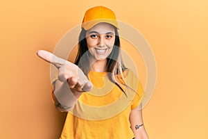 Young hispanic woman wearing delivery uniform and cap smiling friendly offering handshake as greeting and welcoming