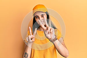 Young hispanic woman wearing delivery uniform and cap smiling looking to the camera showing fingers doing victory sign