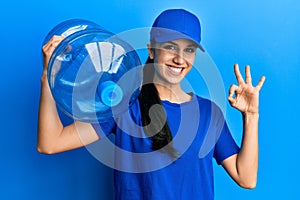 Young hispanic woman wearing delivery uniform holding water carafe doing ok sign with fingers, smiling friendly gesturing