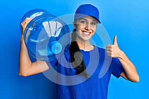 Young hispanic woman wearing delivery uniform holding water carafe smiling happy and positive, thumb up doing excellent and