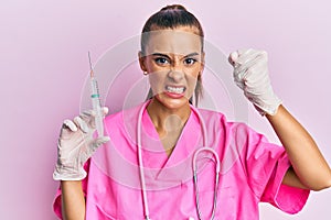 Young hispanic woman wearing doctor stethoscope holding syringe annoyed and frustrated shouting with anger, yelling crazy with