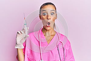 Young hispanic woman wearing doctor stethoscope holding syringe scared and amazed with open mouth for surprise, disbelief face