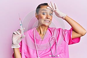 Young hispanic woman wearing doctor stethoscope holding syringe stressed and frustrated with hand on head, surprised and angry