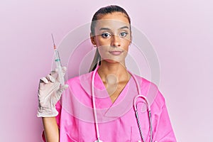 Young hispanic woman wearing doctor stethoscope holding syringe thinking attitude and sober expression looking self confident