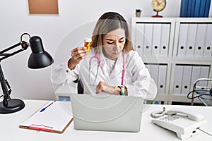 Young hispanic woman wearing doctor uniform holding pills at the clinic checking the time on wrist watch, relaxed and confident