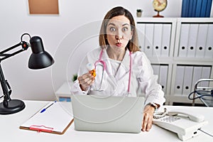 Young hispanic woman wearing doctor uniform holding pills at the clinic puffing cheeks with funny face