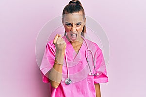 Young hispanic woman wearing doctor uniform and stethoscope angry and mad raising fist frustrated and furious while shouting with