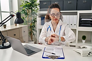 Young hispanic woman wearing doctor uniform and stethoscope checking the time on wrist watch, relaxed and confident