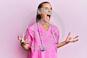 Young hispanic woman wearing doctor uniform and stethoscope crazy and mad shouting and yelling with aggressive expression and arms