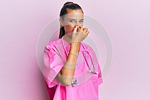 Young hispanic woman wearing doctor uniform and stethoscope smelling something stinky and disgusting, intolerable smell, holding