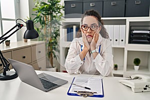 Young hispanic woman wearing doctor uniform and stethoscope tired hands covering face, depression and sadness, upset and irritated