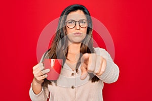 Young hispanic woman wearing glasses drinking a cup of coffee over red background pointing with finger to the camera and to you,