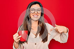 Young hispanic woman wearing glasses drinking a cup of coffee over red background with surprise face pointing finger to himself