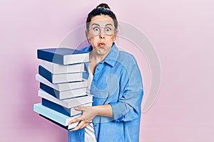 Young hispanic woman wearing glasses and holding books puffing cheeks with funny face