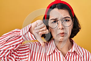 Young hispanic woman wearing glasses over yellow background with angry face, negative sign showing dislike with thumbs down,