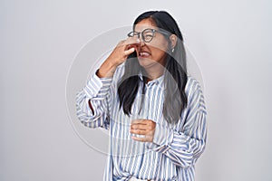 Young hispanic woman wearing glasses smelling something stinky and disgusting, intolerable smell, holding breath with fingers on