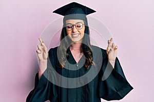 Young hispanic woman wearing graduation cap and ceremony robe gesturing finger crossed smiling with hope and eyes closed