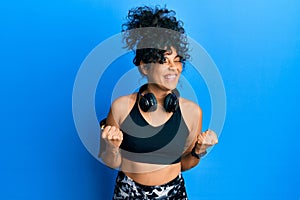 Young hispanic woman wearing gym clothes and using headphones excited for success with arms raised and eyes closed celebrating