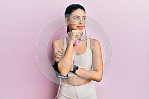 Young hispanic woman wearing gym clothes and using headphones with hand on chin thinking about question, pensive expression